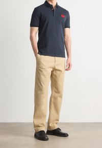 Granatowa koszulka polo z krótkim rękawem i czerwoną naszywką, zestawiona z beżowymi spodniami i czarnymi sznurowanymi butami na neutralnym tle.