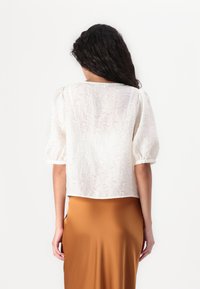 Blusa bianca con texture e maniche a sbuffo, dotata di scollo rotondo. Abbinata a una gonna liscia color caramello.