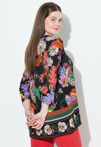 Schwarze Tunika mit lebhaften Blumen- und Blättermustern in Rot, Lila und Grün. Mit Dreiviertelärmeln und rundem Ausschnitt.