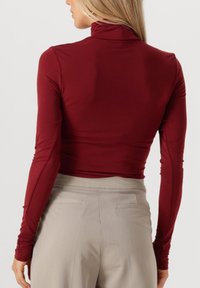 Bordeaux lange mouwen top met een hoge kraag, gemaakt van een gladde stof. Gecombineerd met lichtgrijze broeken met achterzakken.