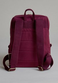 Mochila de tela burdeos con correas ajustables, un asa en la parte superior y detalle de costura horizontal en la parte posterior. Accesorios en tono dorado.