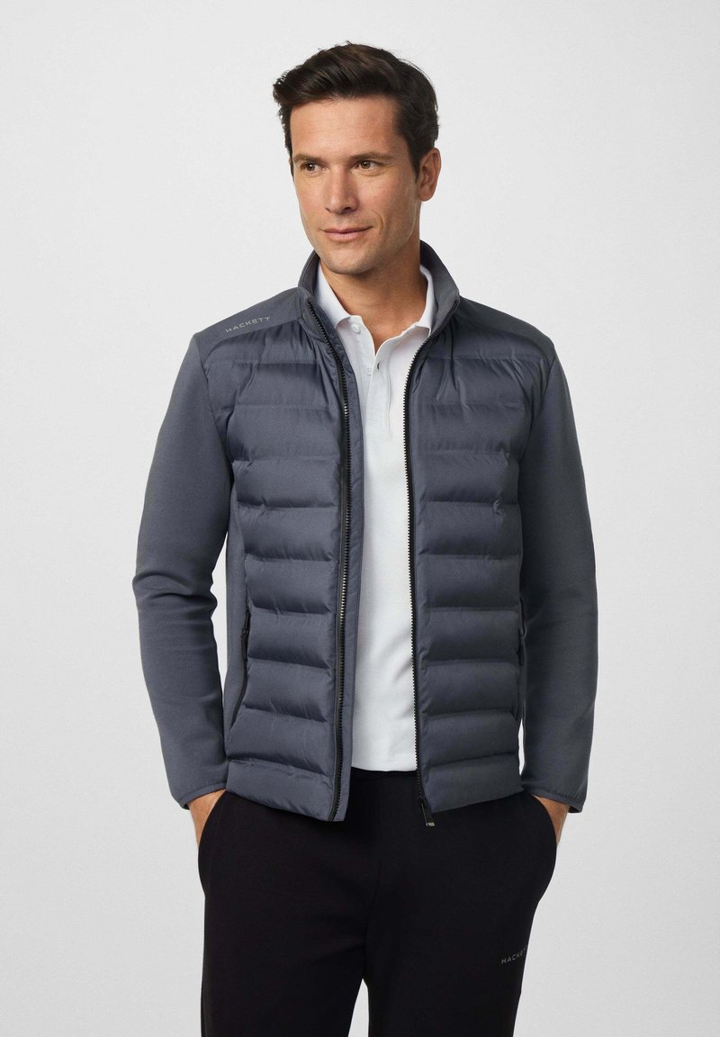 Hombre vestido con una chaqueta de punto en color marino sobre una camiseta polo blanca, de pie con las manos en los bolsillos, mirando ligeramente hacia un lado, sobre un fondo liso.