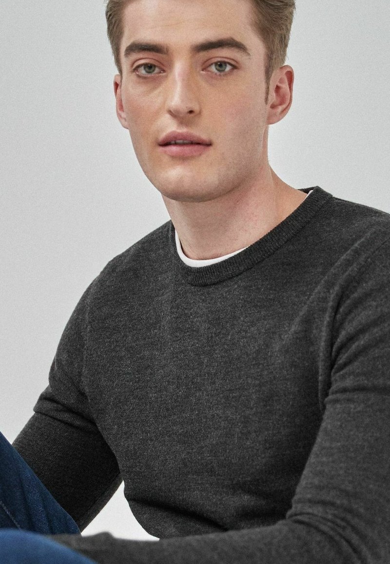 Pulover gri închis cu guler rotund, tricotat texturat, purtat peste un tricou alb. Modelul are părul castaniu deschis și adoptă un stil minimalist.