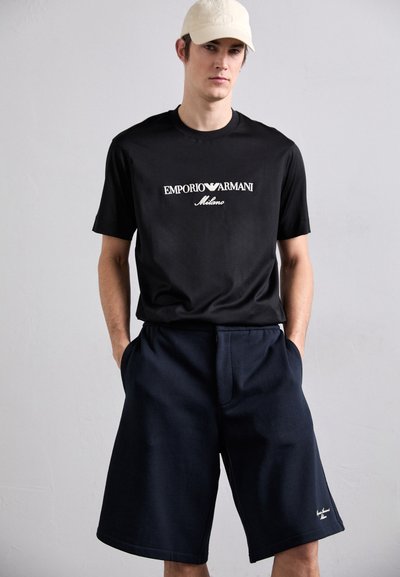 Juodas medvilnės marškinėliai su balta "Emporio Armani Milano" logo, derinami su tamsiai mėlynomis šortais iki kelių, turinčiais kišenes ir minimalų detalizavimą.