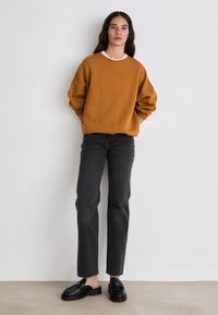 Suéter de gran tamaño color óxido, jeans rectos gris oscuro y zapatos negros sin cordones. Textura suave, diseño simple y acentos mínimos.