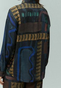 Persona che indossa una camicia oversized scura con motivi geometrici astratti in blu, verde e marrone, fotografata di spalle.