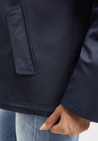 Marineblaue Regenjacke mit glatter Textur, ausgestattet mit einer Pattentasche und einem ordentlich vernähten Ärmelsaum. Eine Hand hält die Jacke fest.