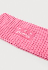 Ροζ πλεκτή κορδέλα κεφαλής με ριγωτή υφή, που διαθέτει τετράγωνο ταμπελάκι με λευκή επιγραφή «TOPH STUDIOS». Μαλακό υλικό, συμπαγής μορφή.