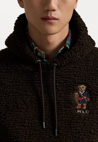 Bruine, gestructureerde hoodie met een kussensurface, voorzien van een geborduurd ontwerp van een beer en koordjes. Een geruit overhemd is zichtbaar onder de kraag.