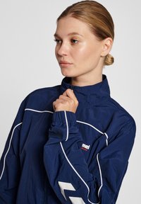 Jonge vrouw met blond haar in een knot, draagt een marineblauwe jas met witte bies en rits, houdt de rits bij haar kin vast.