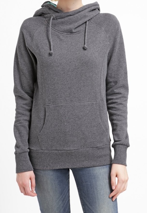 Femme portant un sweat à capuche gris foncé avec une poche kangourou à l'avant et des cordons, associée à un jean bleu, debout devant un fond blanc.