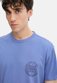 Camiseta de manga corta y cuello redondo en algodón de color azul claro. Presenta un logotipo circular impreso en negro en el lado izquierdo del pecho con texto y acentos de laurel.