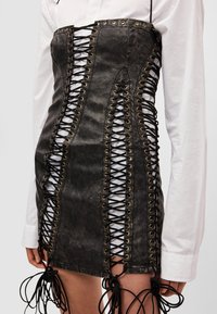 Crna traper haljina-korzet s detaljima od metalnih ringli i vezicama sa strana, s čvorastim naglascima na rubu i struktuiranim krojem.