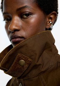 Hnedý kabát s vysokým golierom vyrobený z kordurového materiálu, s detailom gombíka a hladkým povrchom. Detailný záber na tvár.