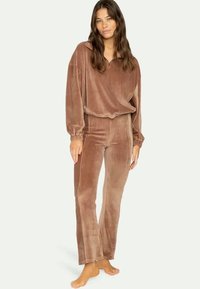 Γυναίκα που στέκεται ξυπόλητη φορώντας ένα καφέ καπιτονέ μπλουζάκι με μακριά μανίκια και αντίστοιχες ψηλόμεσες παντελόνες σε απλή ανοιχτόχρωμη φόντο.