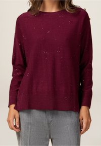 Viininpunainen kevyt neulepusero pyöreällä pääntiellä, jossa on hienovarainen hohto, resorit hihansuissa ja rento istuvuus, yhdistetty harmaisiin housuihin.