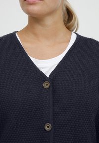 Navy knitted cardigan with a textured pattern, V-neck, and two round brown buttons at the front, worn over a white shirt.