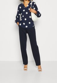 Mujer vestida con pantalones azul marino y una blusa azul marino con puntos blancos, sosteniendo un bolso blanco de hombro y usando sandalias de tacón blancas.