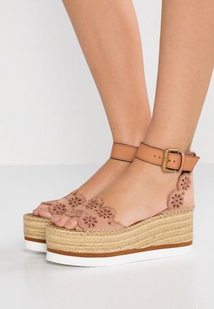 Pieds portant des sandales espadrilles à plateforme beige avec un motif floral découpé et des brides de cheville avec boucles dorées sur un sol blanc.