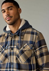 Geruite shirtjack in beige, marineblauw en zwart; heeft een schapenvachtgevoerd capuchon, twee borstzakken en knoopsluitingen.