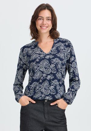 Joven mujer sonriendo, lleva una camiseta de manga larga azul marino con estampados florales blancos y vaqueros oscuros, con las manos en los bolsillos.