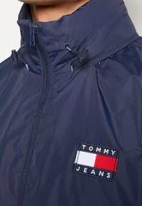 Rompevientos navy con cuello alto, cierre de cremallera y cordones ajustables. Presenta un parche de marca con el logo "TOMMY JEANS" en blanco y rojo.