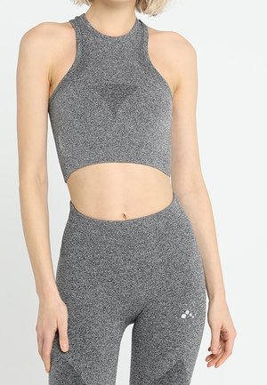 Kobieta ubrana w dopasowany szary bezrękawnik krótki top i legginsy z wysokim stanem z małym białym logo na udzie na jednolitym tle.