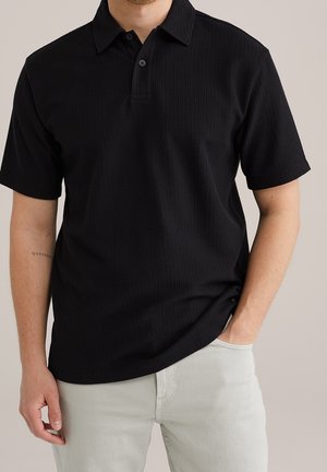 Homme portant un polo côtelé noir à manches courtes et un pantalon clair, une main dans la poche et un tatouage visible sur l’avant-bras gauche.