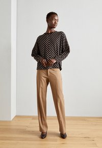 Black and white striped sweater with a loose fit, paired with tan tailored trousers and dark pointed heels. Wooden floor, minimal background.