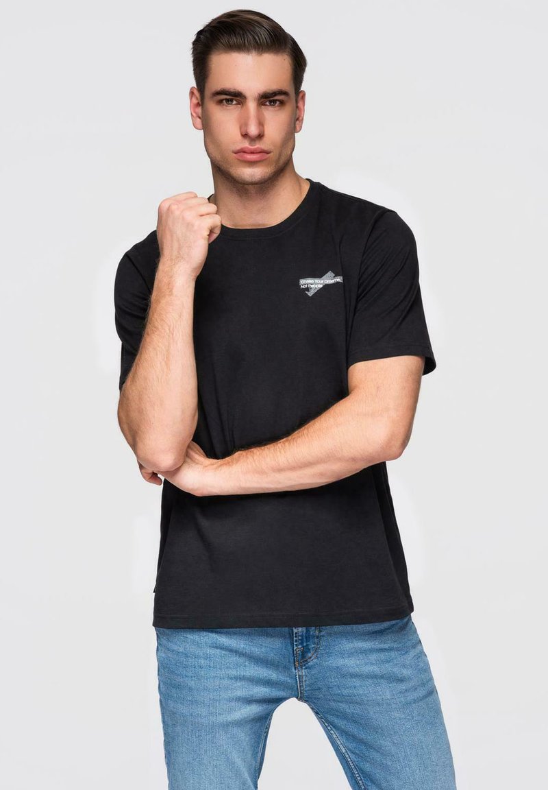 Jeune homme aux cheveux plaqués en arrière, portant un t-shirt noir et un jean bleu, posant avec un poing levé et l'autre bras croisé.