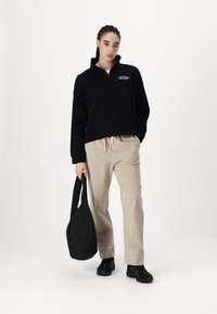 Sudadera negra con cremallera y dobladillo acanalado, pantalones de pana beige y un bolso tote negro acolchado. El modelo lleva zapatillas negras.