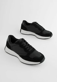 Schwarze Sneakers mit einer Mischung aus glatten und strukturierten Materialien, gepolstertem Schaft, Schnürdesign und einer kontrastierenden weißen Zwischensohle.