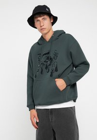 Young man wearing a dark green hoodie with a teardrop eye graphic and black bucket hat, standing with one hand in the hoodie pocket.