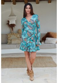 Turkis omslagkjole med paisley-mønster, flænset kant og lange ærmer. Kombineret med beige fransede ankelstøvler. Naturlig baggrund.