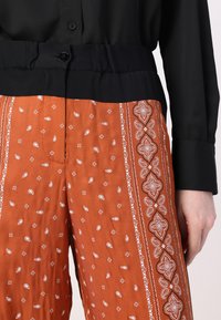 Svart bluse med knappdetaljer, kombinert med høytlivsbukser i rustfarge med mønster av hvite paisley- og geometriske design.