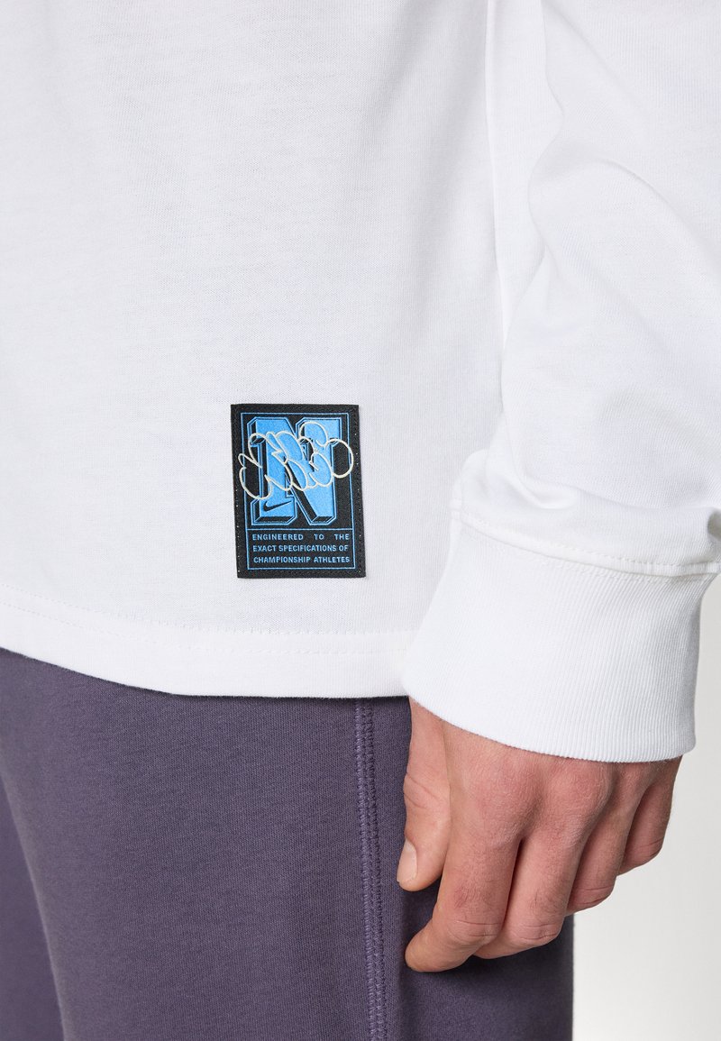 Chemise blanche à manches longues avec une étiquette bleue et noire présentant un design stylisé en "N". Associée à un pantalon de survêtement violet affichant un détail de couture.