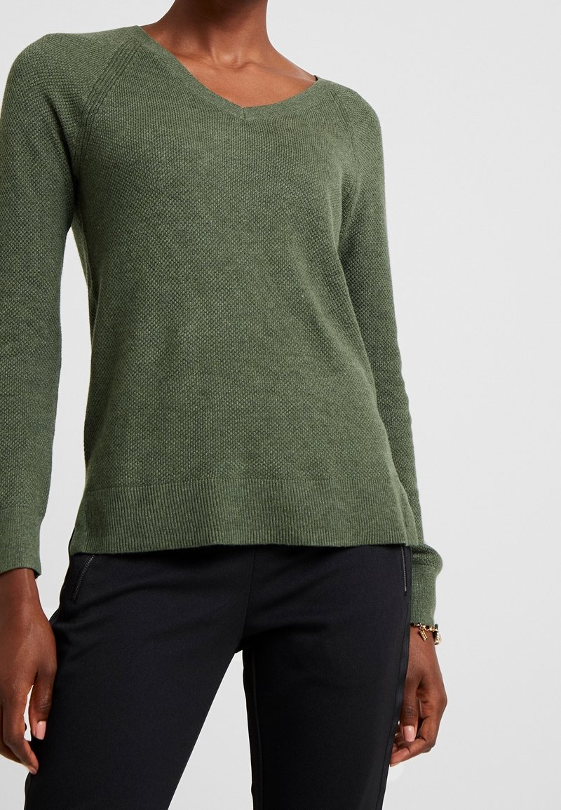 Maglione verde testurizzato con scollo a V, maniche lunghe e orlo a coste. Abbinato a pantaloni neri, che mostrano un outfit casual.