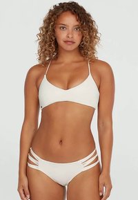 Kvinna med lockigt hår som bär en vit bikinitopp och -botten med remmar, stående mot en enkel ljus bakgrund.