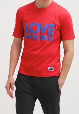 Homme portant un t-shirt rouge à manches courtes avec un grand texte bleu « LOVE MOSCHINO » et un pantalon noir, main dans la poche.