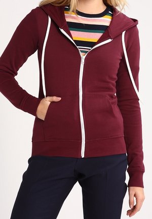 Kobieta nosząca bordową bluzę na zamek z białymi sznurkami, pod nią wielokolorową koszulę w paski oraz ciemnogranatowe spodnie.