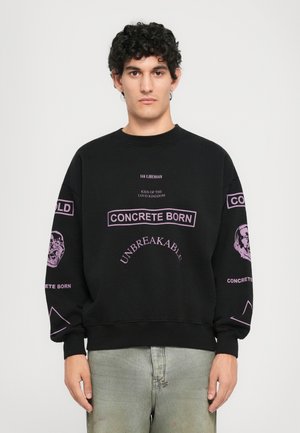 Jonge man met krullend haar die een zwart oversized sweatshirt draagt met paarse tekst en grafische elementen, gecombineerd met licht gewassen jeans, staande voor een witte achtergrond.