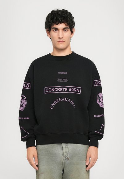 Joven con cabello rizado vestido con un sudadera negra de gran tamaño con texto y gráficos morados, combinado con jeans de lavado claro, de pie frente a un fondo blanco.