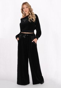 Crop top in velluto nero con design off-shoulder, presenta dettagli a forma di cuore con borchie; abbinato a pantaloni a gamba larga con tasche borchiate.