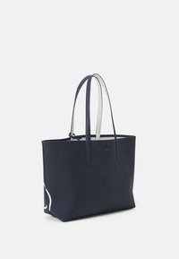 Sac fourre-tout bleu marine en matière texturée, doté de longues poignées et d'un logo discret, avec des accents blancs sur les côtés.