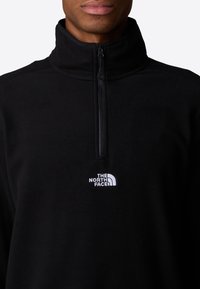 Schwarzer Fleecepullover mit einem halben Reißverschluss am Kragen und weißem, gesticktem Logo auf der Brust. Glatte Textur und klassisches Design.