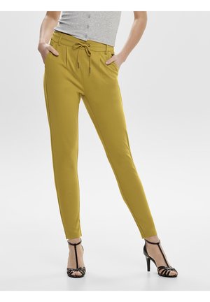 Kobieta ubrana w wąskie musztardowożółte spodnie z wiązanym pasem i czarne sandały na wysokim obcasie, stojąca z rękami w kieszeniach.