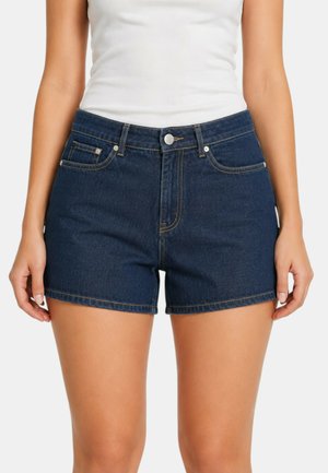Donna che indossa shorts di jeans blu scuro e un top bianco senza maniche, ritratta dalla vita a metà coscia su uno sfondo semplice.