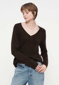 Ženska s kratkimi rjavimi lasmi, oblečena v temnorjavo pulover z V-izrezom, svetloblu jeans in tanek zlat verižico, pozira pred belo ozadjem.