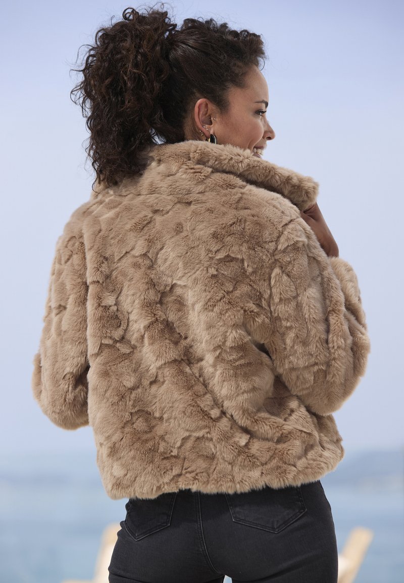 Lažna krznena jakna v svetlo rjavi barvi z mehkim, teksturiranim površjem, velikim ovratnikom in skrajšanim dizajnom, v kombinaciji s temnimi kavbojkami.