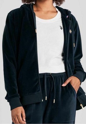 Person iført en mørkeblå velour lynlås-hættetrøje og matchende bukser, med en hvid t-shirt med en lille sort logo på brystet.